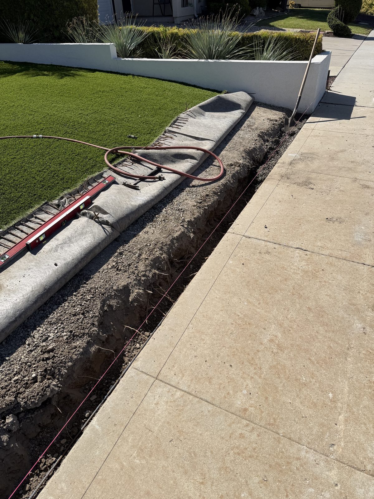 A B&M trench excavated to depth along a walkway, with compacted gravel base, geotextile fabric, a taut string line and a level